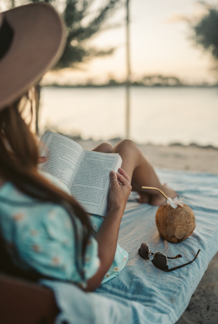 Image of a woman lounging on the beach reading a book with a coconut drink.